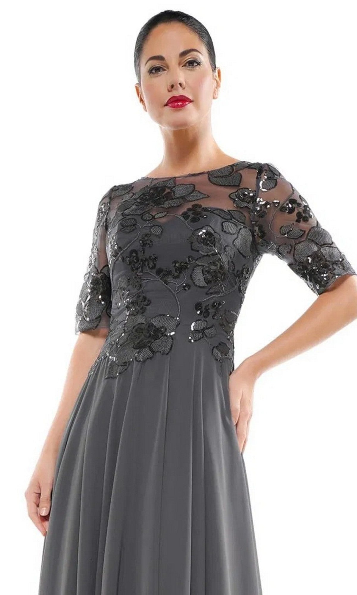A woman models the Marsoni M286 Long Formal Dress—dark gray with a sheer floral embroidered bodice and short sleeves—against a white background.